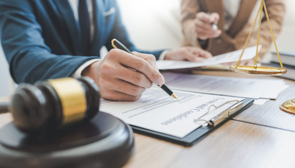 Close-up of a lawyer in a suit signing legal documents at a desk, with a gavel and scales of justice in the foreground.