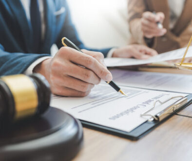 Close-up of a lawyer in a suit signing legal documents at a desk, with a gavel and scales of justice in the foreground.