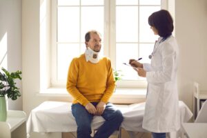 A man wearing a neck brace sits on an examination table while a doctor takes notes, representing medical treatment after a personal injury.