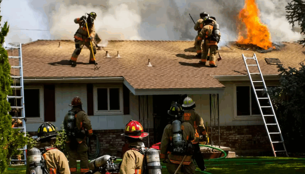 Firefighters battling a house fire while flames shoot from the roof and smoke fills the air